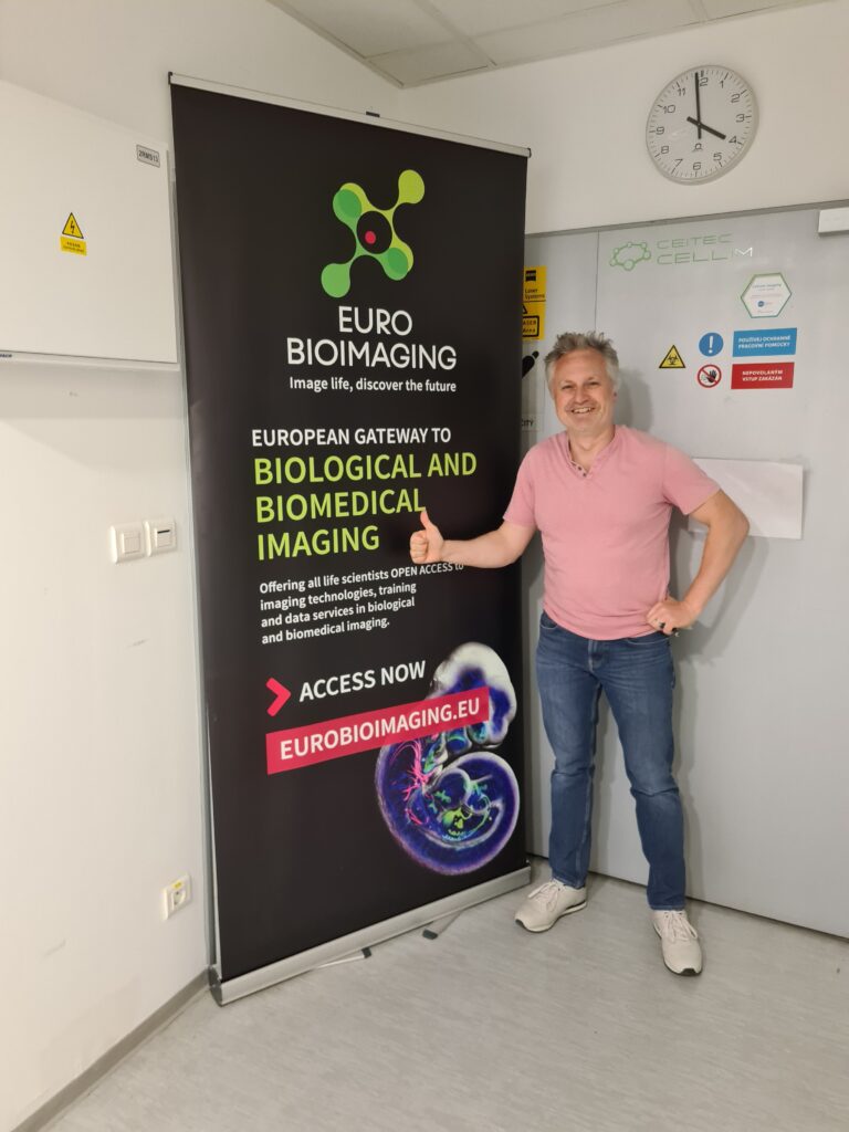 Milan Esner, Deputy Head of the Brno Node and Representative of microscopic imaging within node in front of a Euro-BioImaging poster in the facility.
